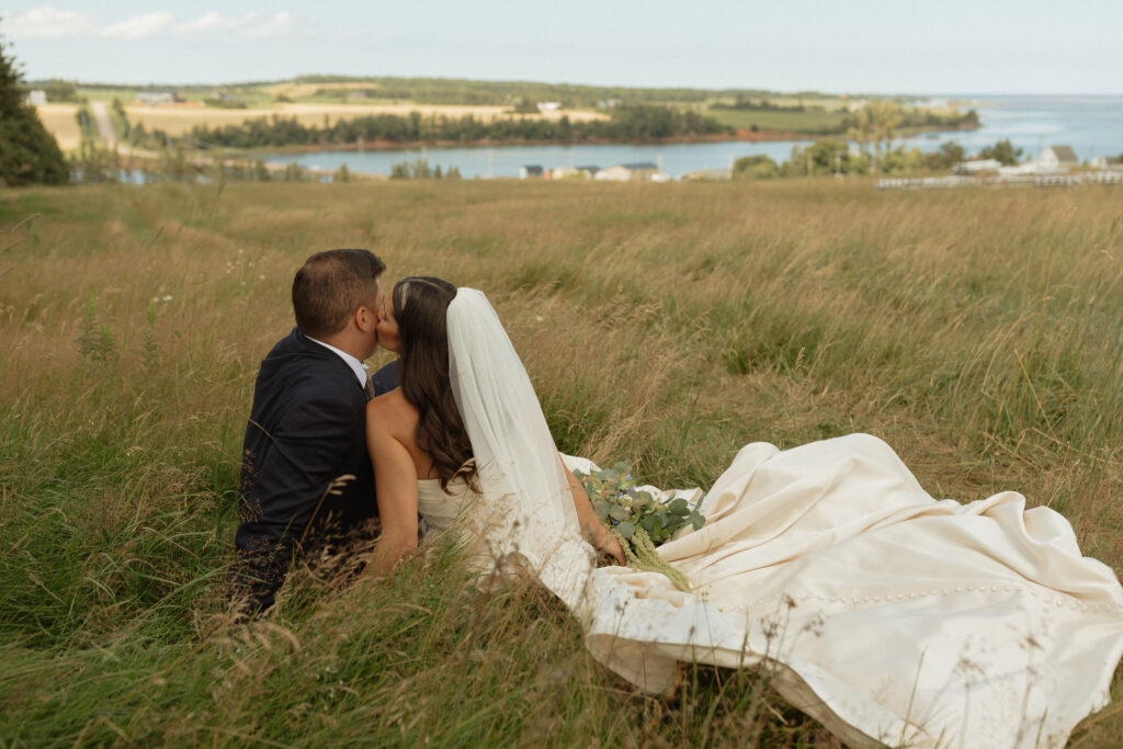 couple sitting in long grass in wedding dress and tux looking out at the view of the ocean and cliffs