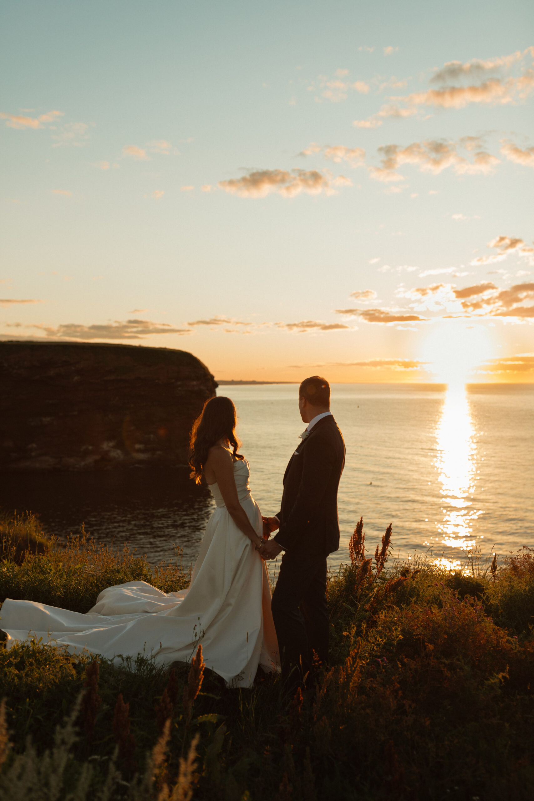 bride and groom looking out at sunset over the ocean while holding hands