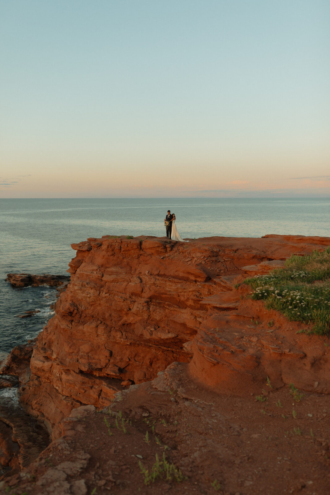 far away photo of bride hugging groom from behind on cliffs at sunset