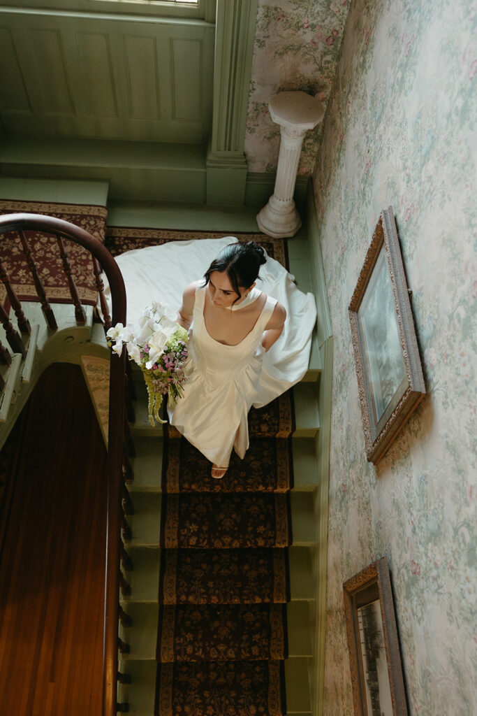 bride walking down spiral staircase holding bouquet while dress trails behind her 