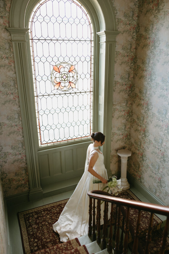bride walking down the staircase, overlooking vintage arch window with stained glass