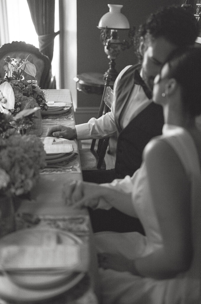 black and white film photo of bride and groom holding hands at wedding dinner