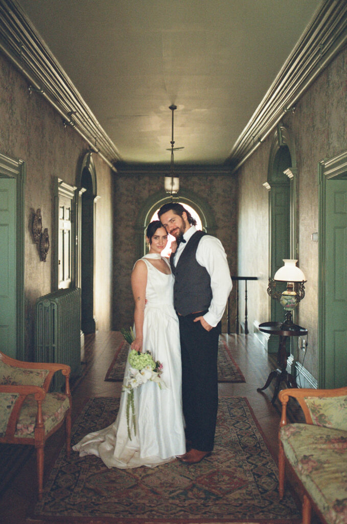 film photo of bride and groom in hallway of vintage hotel posing for the camera, bouquet in hand.